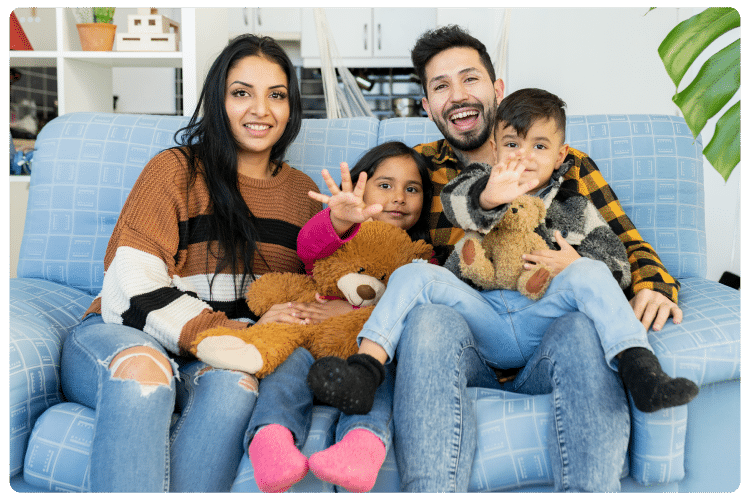 Happy Hispanic family sitting together on a couch at home smiling and waving, representing compassionate family healthcare at NAG Clinics in Pasadena and Texas City, Texas. NAG Clinics provides pediatric care, family medicine, annual wellness exams, asthma treatment, weight loss programs, STI testing, mental health support, LGBTQ+ friendly healthcare, and primary care services for diverse families and the local community.